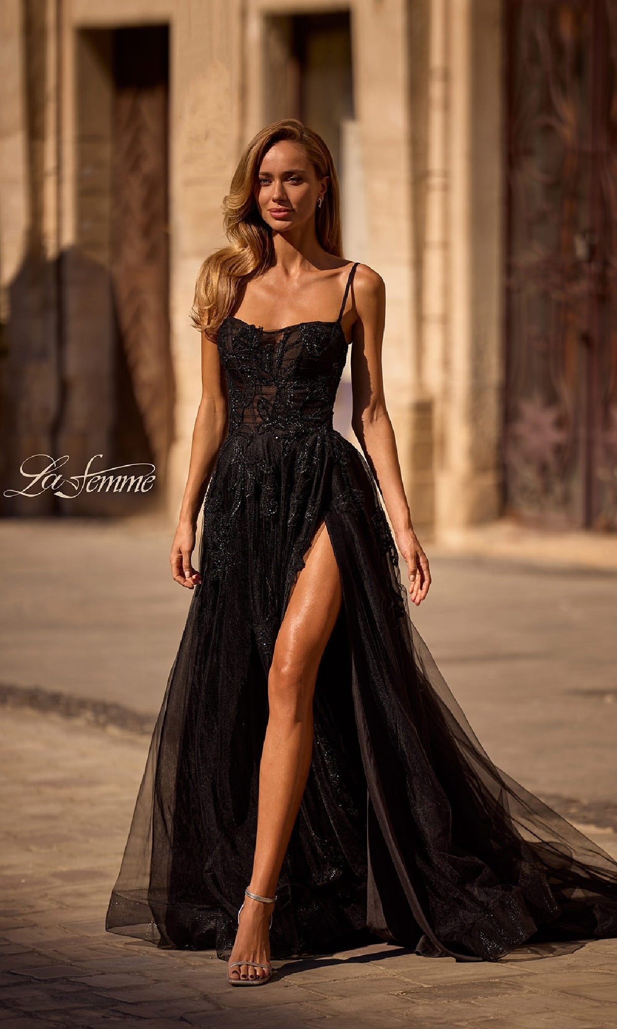 A woman stands outdoors on a sunlit stone street, her hair in loose waves, wearing the Long Black Glitter Prom Dress: La Femme 32781—a sleeveless gown with a high slit.